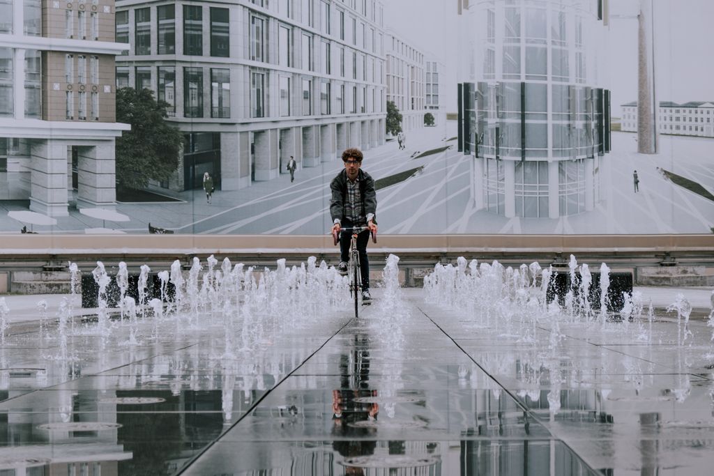 A man cycles through an urban area with modern architecture and reflecting fountains on wet pavement.